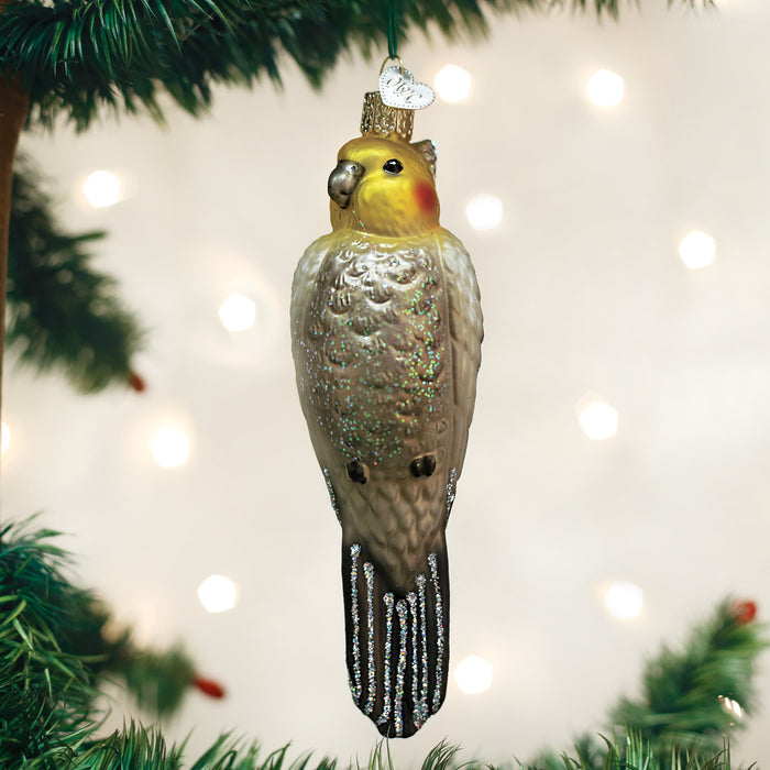 A bird-shaped Christmas tree ornament resembling a cockatiel, with a yellow head, rosy cheeks, silver glittery accents, and a small gold crown, hangs on a green branch against a soft, light-filled background.