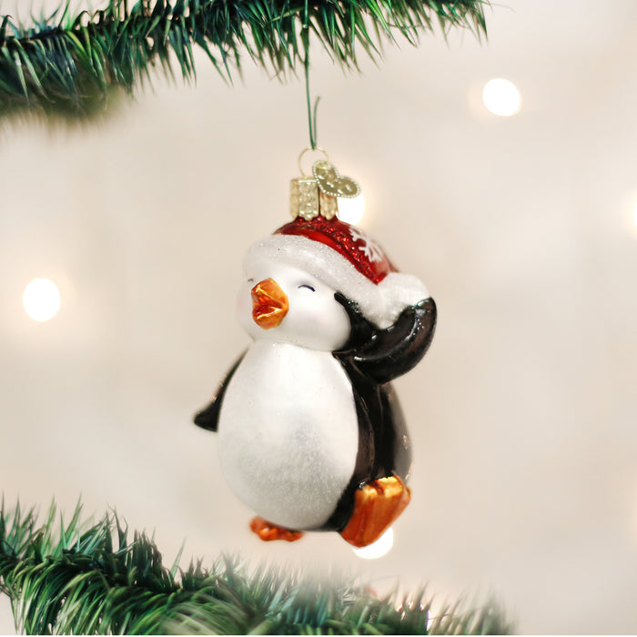 A glass penguin ornament wearing a red Santa hat hangs from a green Christmas tree branch with soft, glowing lights in the background.