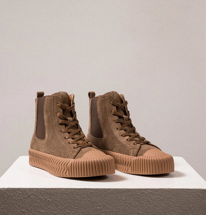 The DANIELLA LEHAVI - OSAKA HIGH TOP SNEAKER features brown suede, tan ribbed rubber soles, and side elastic panels—a stylish and comfy choice from DANIELLA LEHAVI, shown on a white pedestal against a neutral background.