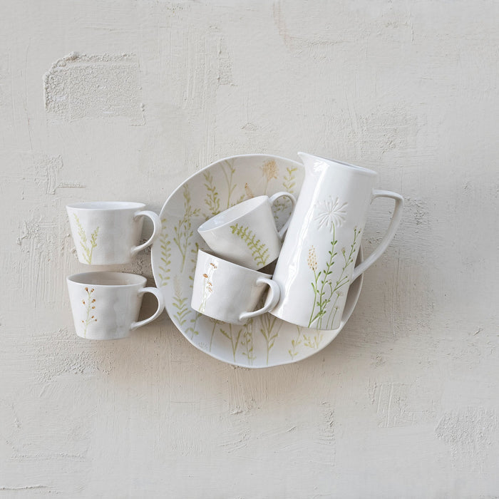 The CREATIVE COOP DEBOSSED STONEWARE PITCHER WITH BOTANICAL PATTERN features a cream finish, handle, spout, and an embossed and painted floral design with green stems and white flowers on one side.
