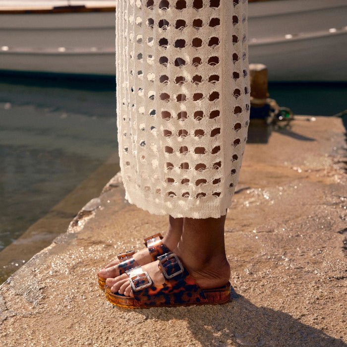 The DOLCE VITA - JULIO SANDAL IN TORTOISE is a jelly slide with a tortoiseshell pattern, two large buckled straps, and a chunky sole, shown against a plain light gray background.