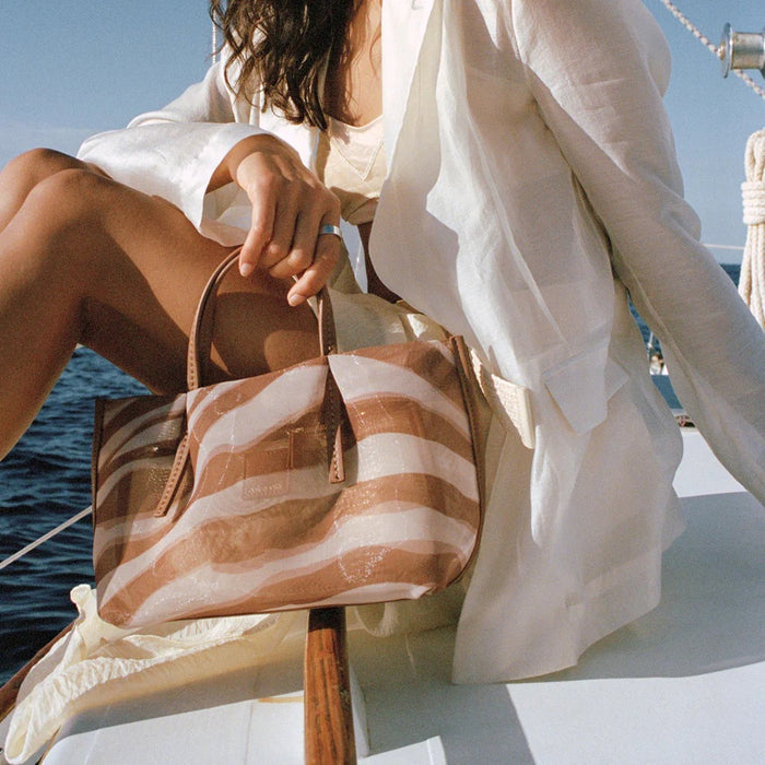 A woman in a light blazer and dress sits on a boat, sunlight highlighting her look and the DOLCE VITA - SELENE ZEBRA MESH BAG resting on her knee. The bright, relaxed scene showcases the stylish bag from DOLCE VITA.