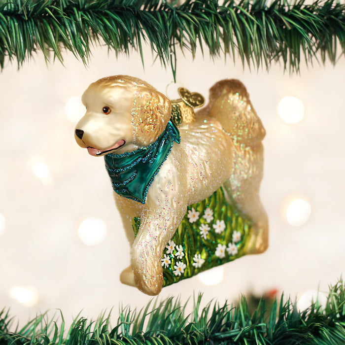 A glittery dog-shaped Christmas ornament wearing a teal scarf hangs from a green Christmas tree branch, with twinkling white lights and a blurred background.
