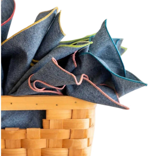 A woven wooden basket displays DOT AND ARMY Chambray Cocktail Napkins—each gray napkin features a different brightly colored stitched edge—all set against a white background.