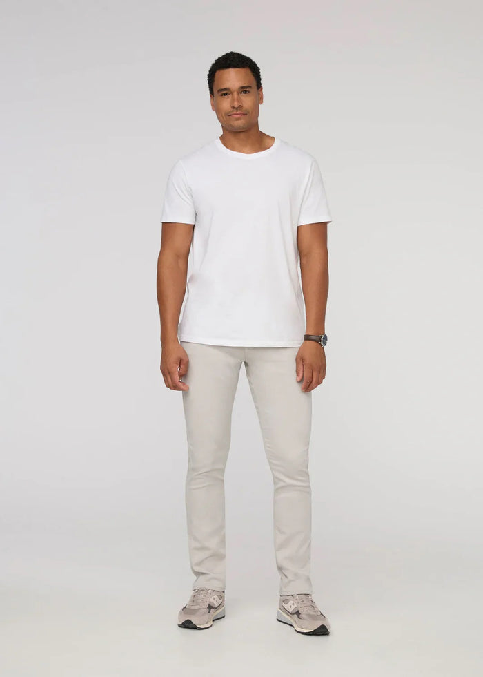 A person stands facing forward on a plain light background, wearing DUER NO SWEAT ATHLETIC TAPER PANTS IN MIST and matching light gray sneakers, feet slightly apart.