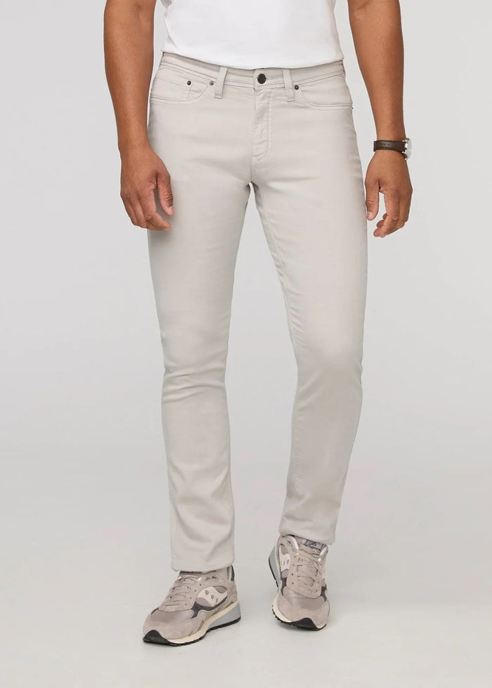 A person stands facing forward on a plain light background, wearing DUER NO SWEAT ATHLETIC TAPER PANTS IN MIST and matching light gray sneakers, feet slightly apart.