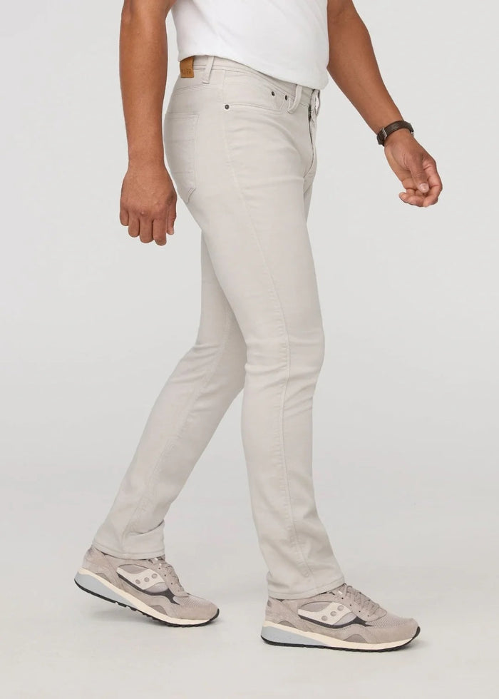 A person stands facing forward on a plain light background, wearing DUER NO SWEAT ATHLETIC TAPER PANTS IN MIST and matching light gray sneakers, feet slightly apart.