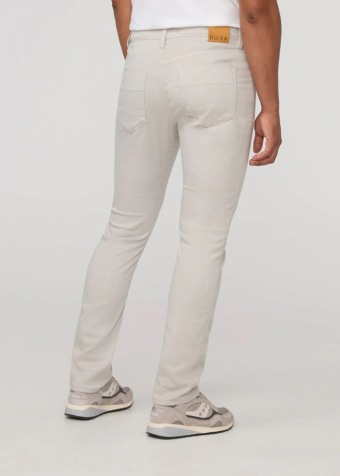 A person stands facing forward on a plain light background, wearing DUER NO SWEAT ATHLETIC TAPER PANTS IN MIST and matching light gray sneakers, feet slightly apart.