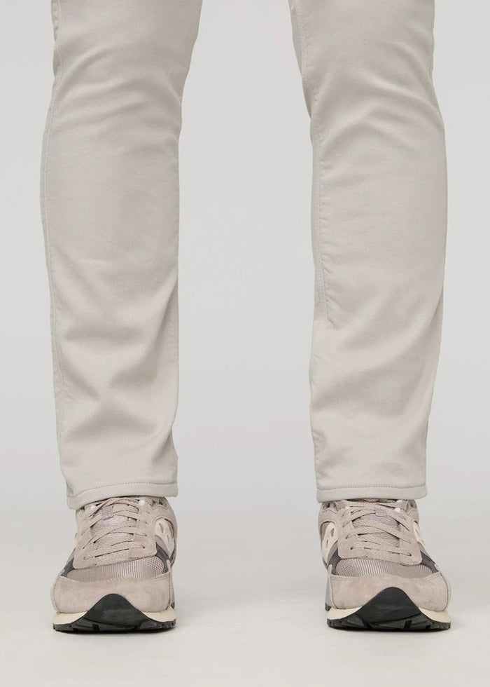 A person stands facing forward on a plain light background, wearing DUER NO SWEAT ATHLETIC TAPER PANTS IN MIST and matching light gray sneakers, feet slightly apart.