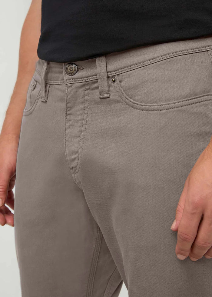 A close-up shows a person wearing DUER No Sweat Athletic Taper Pants in Stone, highlighting the waist, upper legs, stitching, and button detail. The pants are made with No Sweat fabric; the person pairs them with a black shirt.