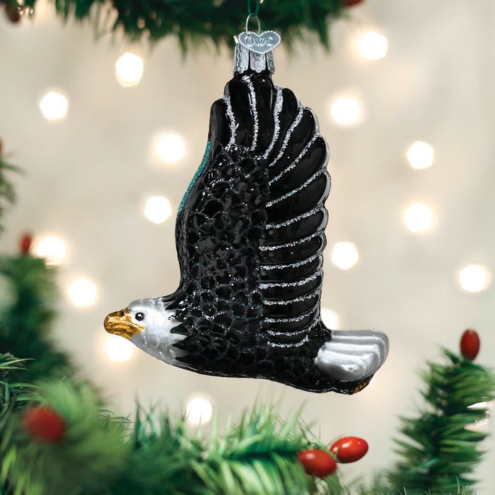 A glass ornament shaped like a flying bald eagle with black wings, a white head, and a gold beak hangs on a green Christmas tree, with blurred white lights in the background.