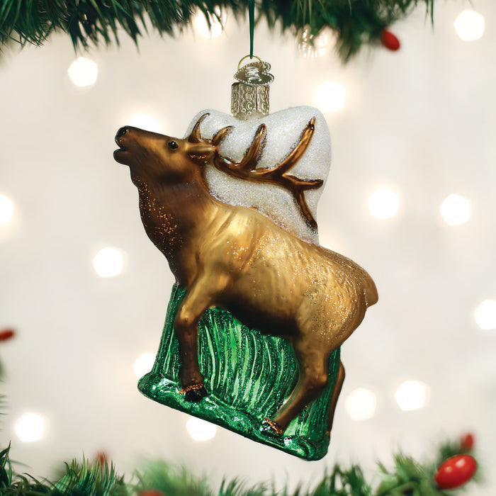 A glass Christmas tree ornament shaped like an elk standing on green grass with snow and antlers, hanging from a tree branch with blurred lights in the background.