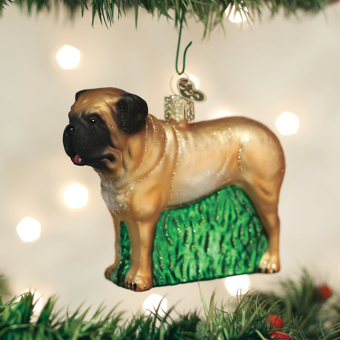A Christmas tree ornament shaped like a light brown dog with a black snout stands on green grass, hanging from a tree branch with blurred white lights in the background.