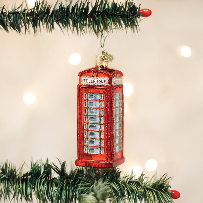 A red glass ornament shaped like a British telephone booth hangs on a Christmas tree branch with blurred white lights in the background.