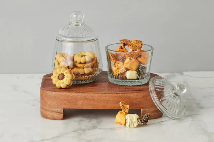 ETUHOME Bistro Glass Biscuit Jars with lids sit on a wooden tray, filled with snacks and orange-wrapped candies. The classic jar design by ETUHOME stands out against the softly blurred background.