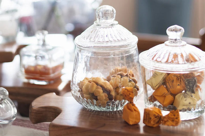 ETUHOME Bistro Glass Biscuit Jars with lids sit on a wooden tray, filled with snacks and orange-wrapped candies. The classic jar design by ETUHOME stands out against the softly blurred background.