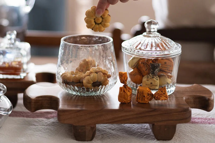 ETUHOME Bistro Glass Biscuit Jars with lids sit on a wooden tray, filled with snacks and orange-wrapped candies. The classic jar design by ETUHOME stands out against the softly blurred background.