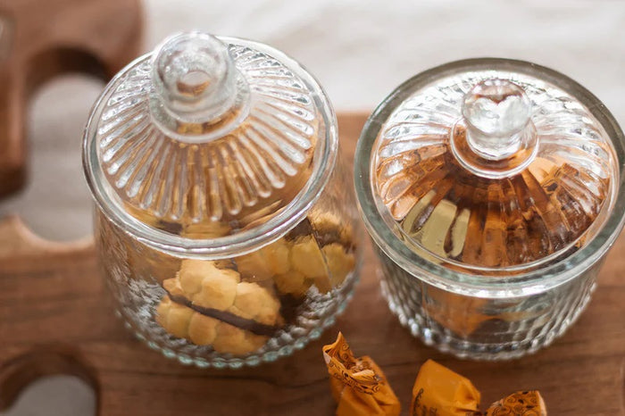 Two ETUHOME Bistro Glass Bon Bon Jars with ribbed lids sit on a wooden surface, one filled with yellow candies and the other nearly empty. Wrapped sweets are scattered nearby, creating a charming ETUHOME display.