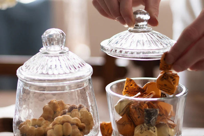 Two ETUHOME Bistro Glass Bon Bon Jars with ribbed lids sit on a wooden surface, one filled with yellow candies and the other nearly empty. Wrapped sweets are scattered nearby, creating a charming ETUHOME display.