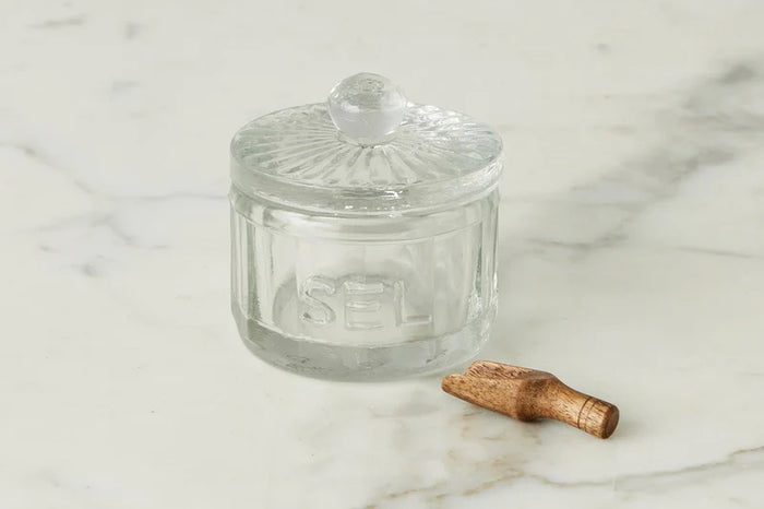 The ETUHOME Bistro Glass Salt Cellar with Scoop sits on a wooden tray atop a marble counter, accompanied by two other glass jars for herbs and sugar. A small wooden scoop from ETUHOME rests in front.