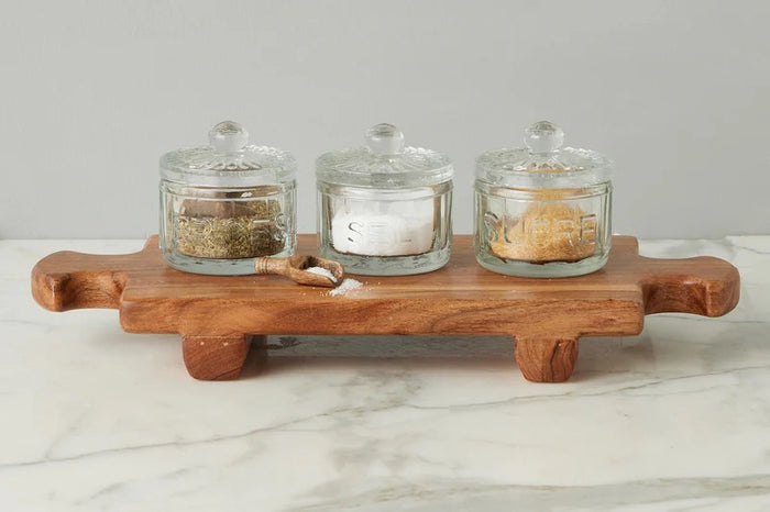 The ETUHOME Bistro Glass Salt Cellar with Scoop sits on a wooden tray atop a marble counter, accompanied by two other glass jars for herbs and sugar. A small wooden scoop from ETUHOME rests in front.