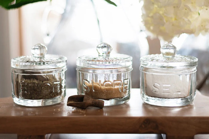 The ETUHOME Bistro Glass Salt Cellar with Scoop sits on a wooden tray atop a marble counter, accompanied by two other glass jars for herbs and sugar. A small wooden scoop from ETUHOME rests in front.
