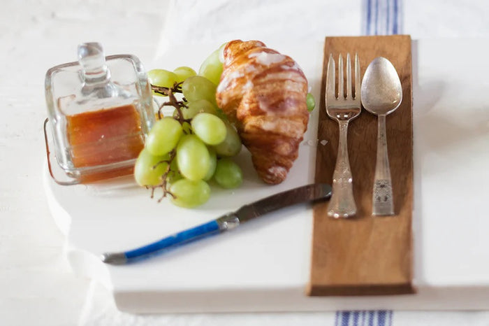 A breakfast setting with a croissant, green grapes, an ETUHOME - BISTRO GLASS SQUARE JAM JAR, a butter knife, fork and spoon on a wooden tray over a white surface with a blue-striped cloth.