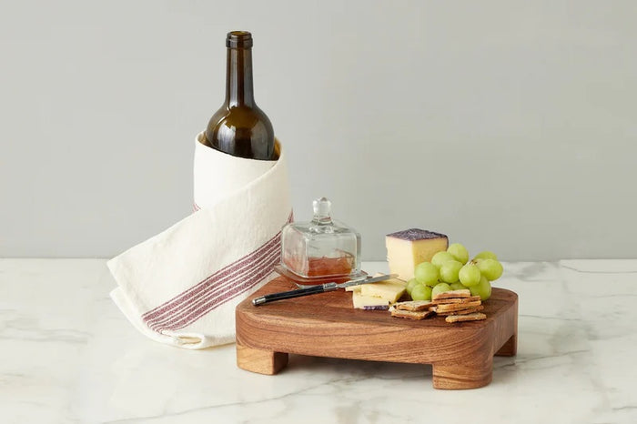 A breakfast setting with a croissant, green grapes, an ETUHOME - BISTRO GLASS SQUARE JAM JAR, a butter knife, fork and spoon on a wooden tray over a white surface with a blue-striped cloth.