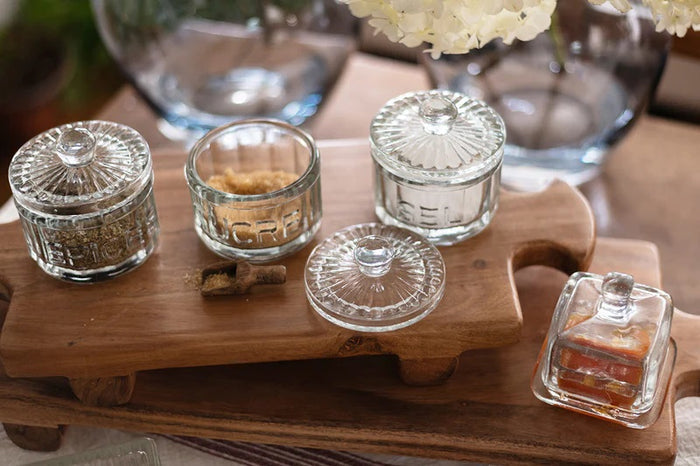 A breakfast setting with a croissant, green grapes, an ETUHOME - BISTRO GLASS SQUARE JAM JAR, a butter knife, fork and spoon on a wooden tray over a white surface with a blue-striped cloth.