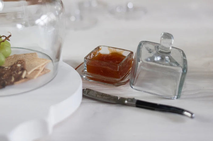 A breakfast setting with a croissant, green grapes, an ETUHOME - BISTRO GLASS SQUARE JAM JAR, a butter knife, fork and spoon on a wooden tray over a white surface with a blue-striped cloth.