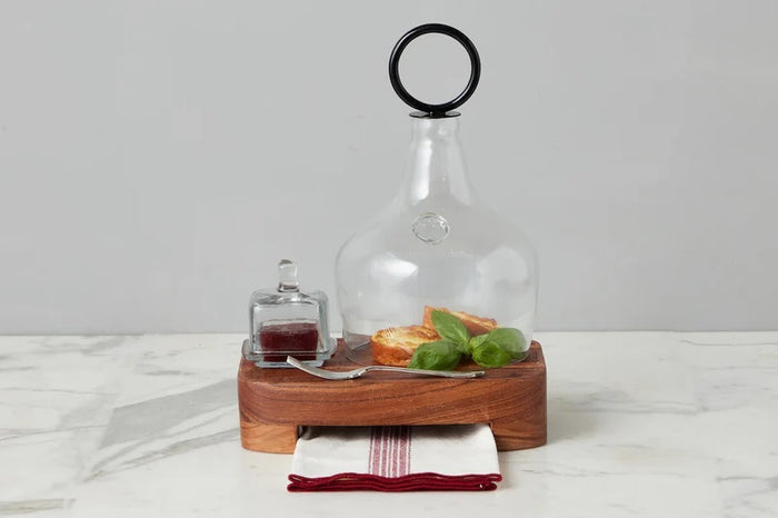 A breakfast setting with a croissant, green grapes, an ETUHOME - BISTRO GLASS SQUARE JAM JAR, a butter knife, fork and spoon on a wooden tray over a white surface with a blue-striped cloth.