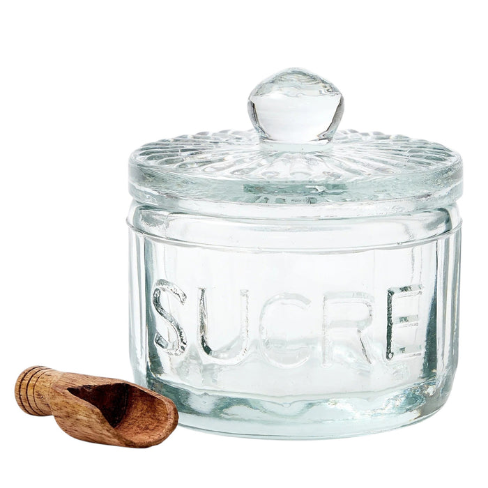The ETUHOME Bistro Glass Sugar Cellar with Scoop by ETUHOME holds brown sugar on a white countertop near a folded white towel with red stripes and a wooden tray, adding classic charm to your kitchen storage collection.