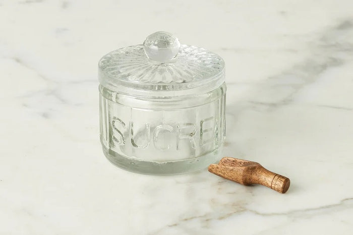 The ETUHOME Bistro Glass Sugar Cellar with Scoop by ETUHOME holds brown sugar on a white countertop near a folded white towel with red stripes and a wooden tray, adding classic charm to your kitchen storage collection.