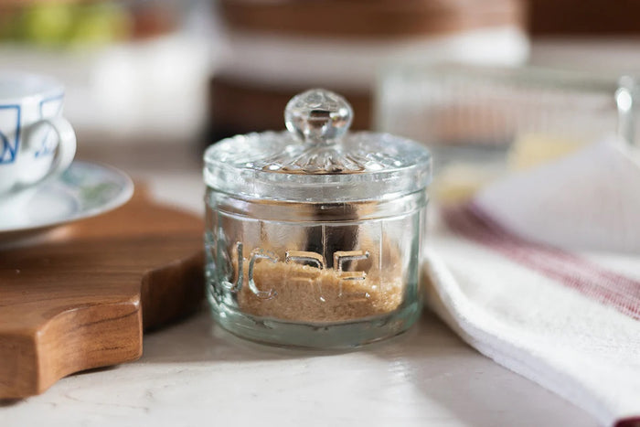 The ETUHOME Bistro Glass Sugar Cellar with Scoop by ETUHOME holds brown sugar on a white countertop near a folded white towel with red stripes and a wooden tray, adding classic charm to your kitchen storage collection.