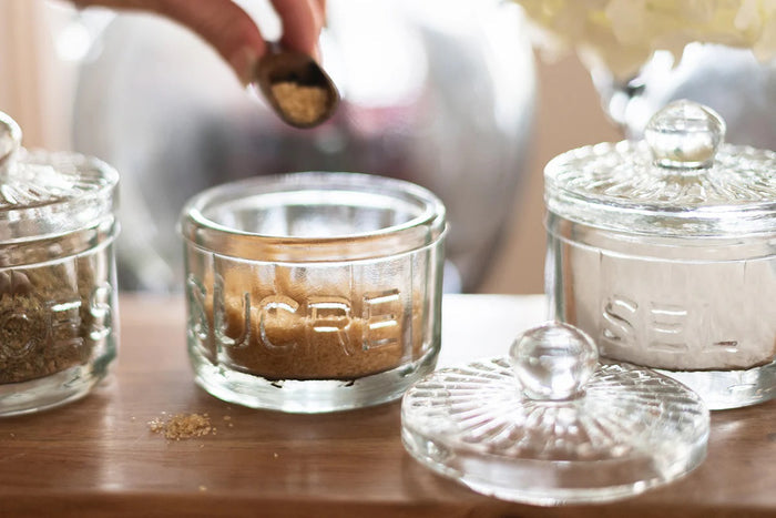 The ETUHOME Bistro Glass Sugar Cellar with Scoop by ETUHOME holds brown sugar on a white countertop near a folded white towel with red stripes and a wooden tray, adding classic charm to your kitchen storage collection.