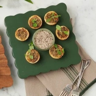 The ETUHOME Green Square Scalloped Cheese Board with Markers displays six savory herb tarts arranged in a circle around a glass dip bowl, resting on a beige napkin with two forks on a marble surface.