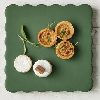 The ETUHOME Green Square Scalloped Cheese Board with Markers displays six savory herb tarts arranged in a circle around a glass dip bowl, resting on a beige napkin with two forks on a marble surface.