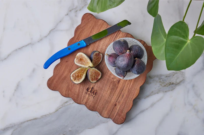 The ETUHOME Natural Square Scalloped Cutting Board, Medium, crafted from durable acacia wood, stands upright against a light gray wall on a white marble surface.
