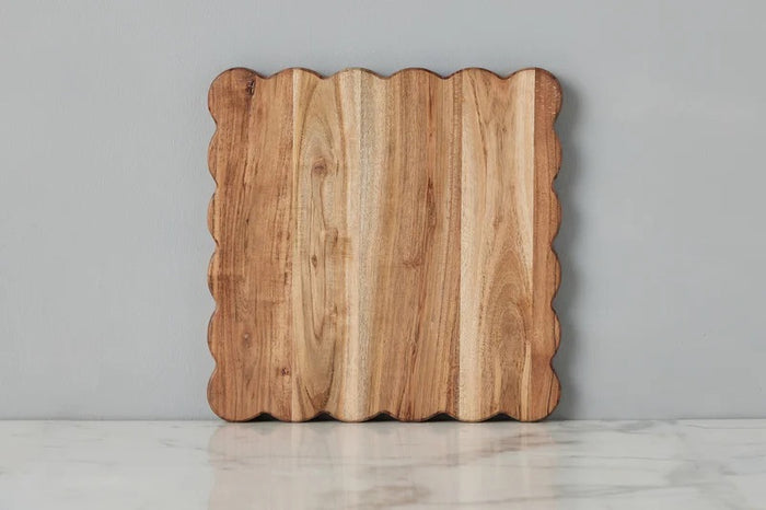 The ETUHOME Natural Square Scalloped Cutting Board, Medium, crafted from durable acacia wood, stands upright against a light gray wall on a white marble surface.