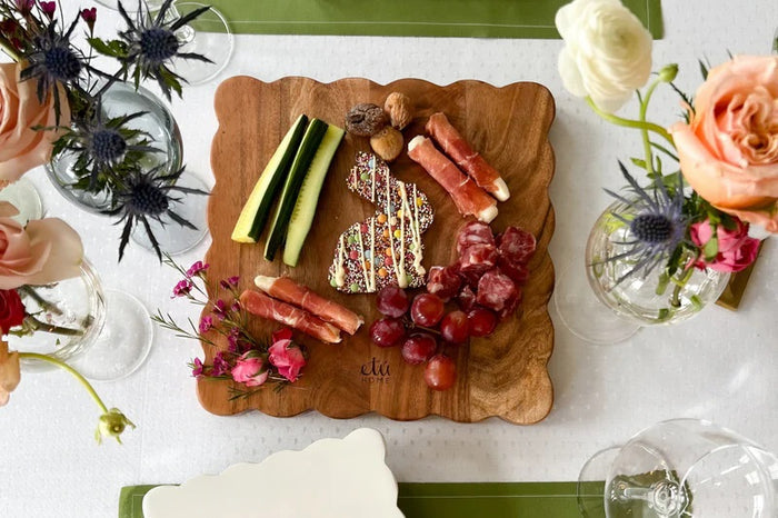 The ETUHOME Natural Square Scalloped Cutting Board, Medium, crafted from durable acacia wood, stands upright against a light gray wall on a white marble surface.