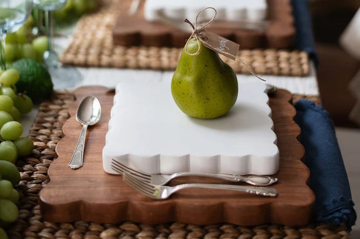 The ETUHOME Natural Square Scalloped Cutting Board, Medium, crafted from durable acacia wood, stands upright against a light gray wall on a white marble surface.