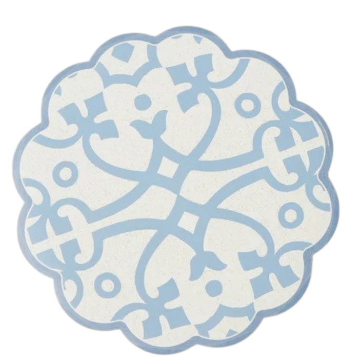 The ETUHOME Round Scalloped Placemat in Denim, featuring a blue and white geometric floral pattern, is displayed on a light wooden surface with green plant leaves visible in the upper right corner.