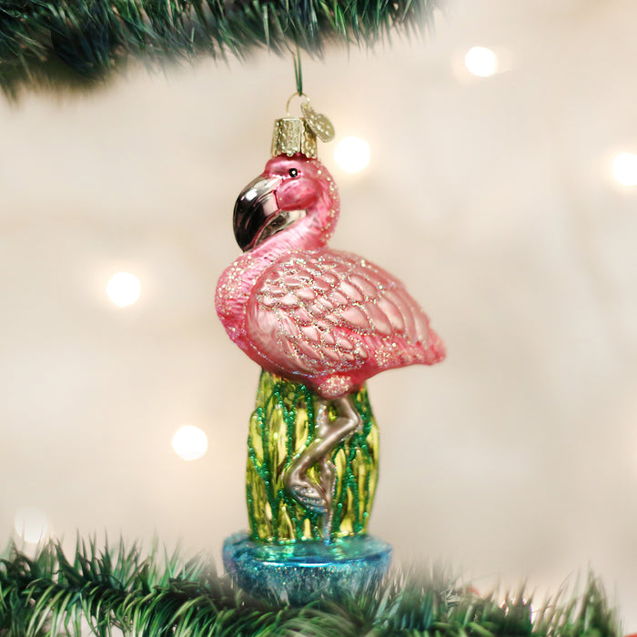 A pink flamingo ornament with glitter details hangs from a Christmas tree branch, surrounded by green pine needles and blurred white holiday lights in the background.