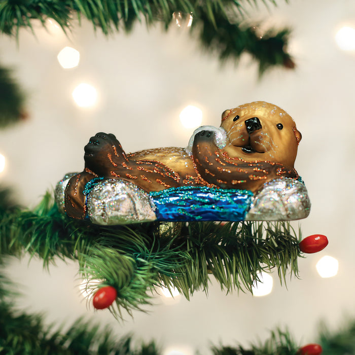 A Christmas tree ornament shaped like an otter floating on its back in blue water, surrounded by green pine needles and red berries, with blurred white lights in the background.