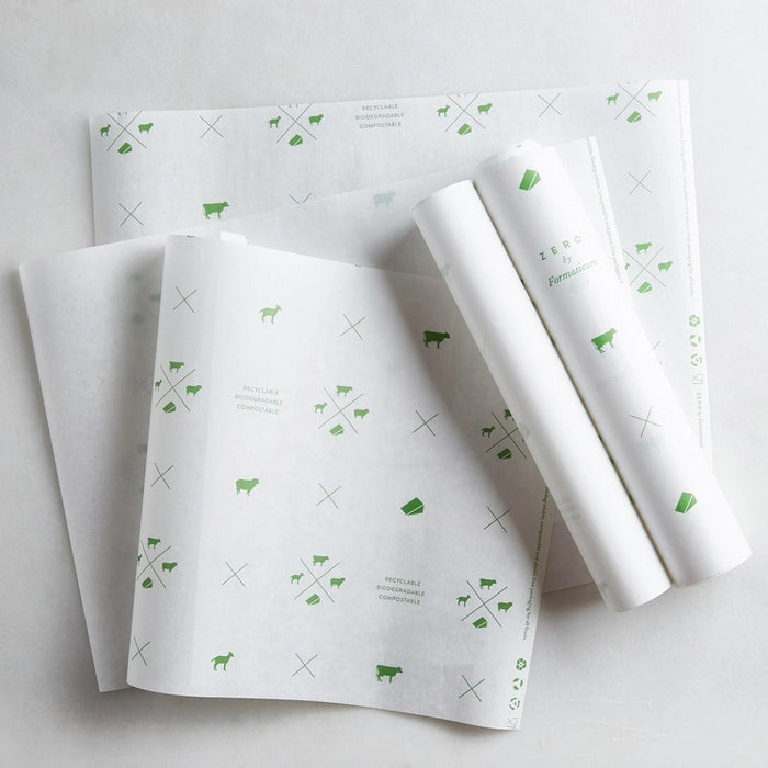 A hand holds a sheet of FORMATICUM - ZERO CHEESE STORAGE PAPER with green illustrations, next to a brown FORMATICUM box. The partially unrolled cheese storage paper is ideal for keeping cheese fresh at home.