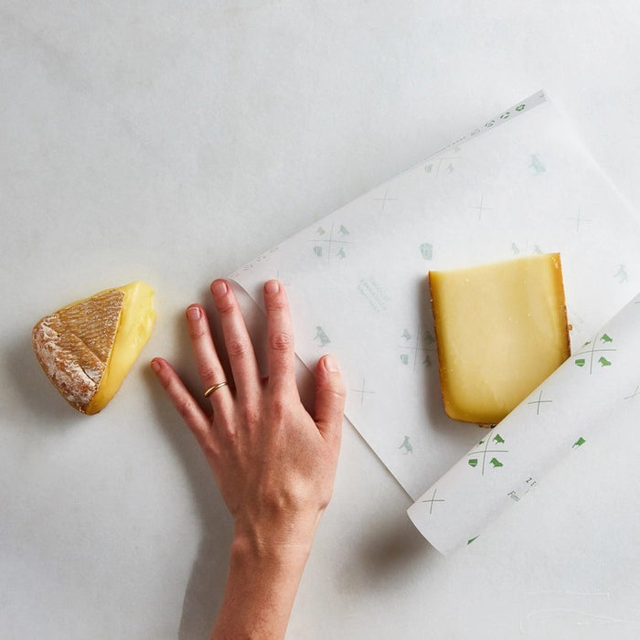 A hand holds a sheet of FORMATICUM - ZERO CHEESE STORAGE PAPER with green illustrations, next to a brown FORMATICUM box. The partially unrolled cheese storage paper is ideal for keeping cheese fresh at home.