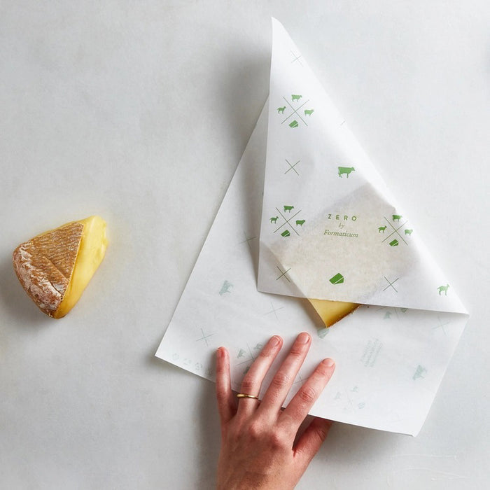 A hand holds a sheet of FORMATICUM - ZERO CHEESE STORAGE PAPER with green illustrations, next to a brown FORMATICUM box. The partially unrolled cheese storage paper is ideal for keeping cheese fresh at home.