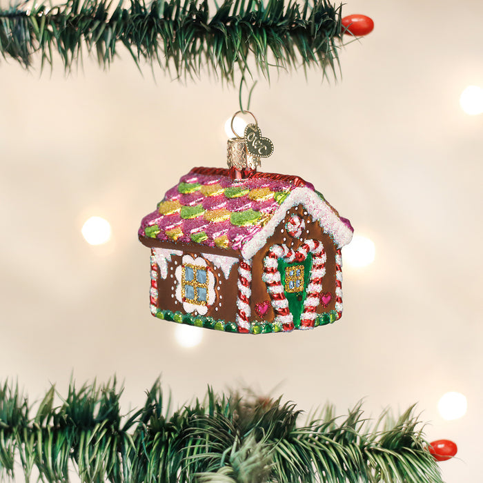 A colorful gingerbread house ornament with candy decorations hangs from a Christmas tree branch, with blurred white holiday lights in the background.
