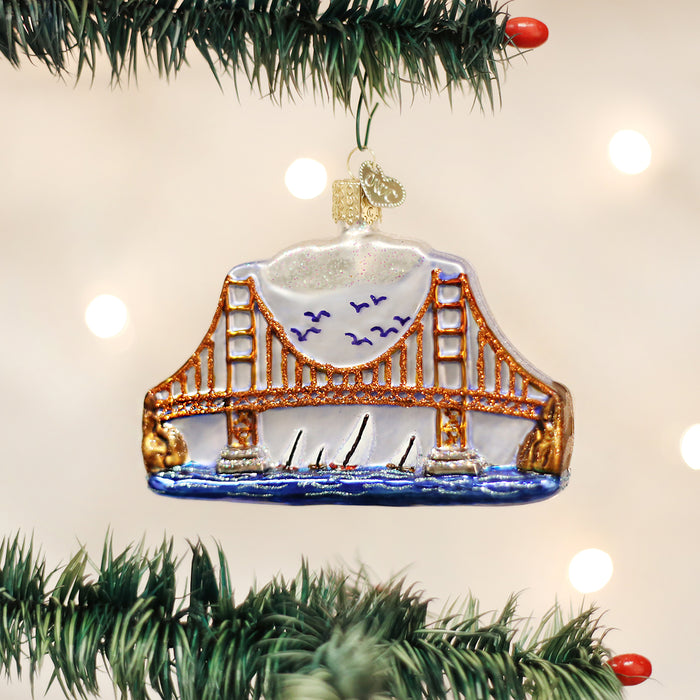 A glass ornament shaped like the Golden Gate Bridge hangs on a Christmas tree branch, with blurred lights in the background.