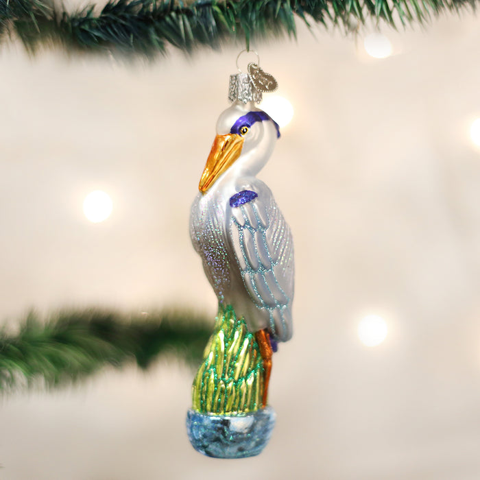 A glass Christmas tree ornament shaped like a heron with blue, white, and silver feathers, orange beak and legs, perched on green reeds, hanging from a pine branch.